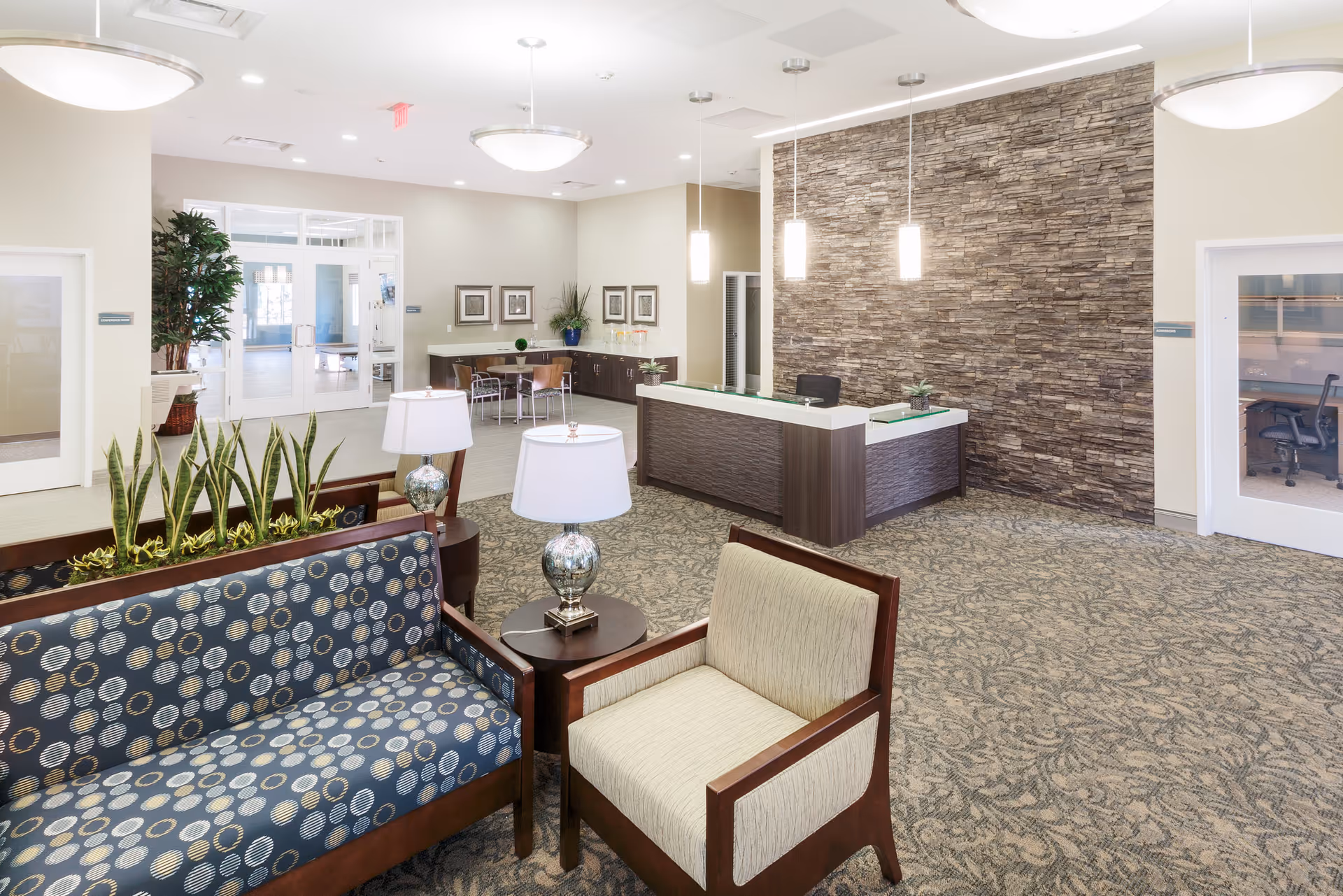 A spacious and well-lit reception area with a modern front desk against a stone accent wall. The room features patterned carpet, comfortable seating including a blue patterned sofa and a beige armchair, two table lamps, and several plants. In the background, there is a small table with chairs and framed artwork on the wall, as well as glass doors leading to other rooms.
