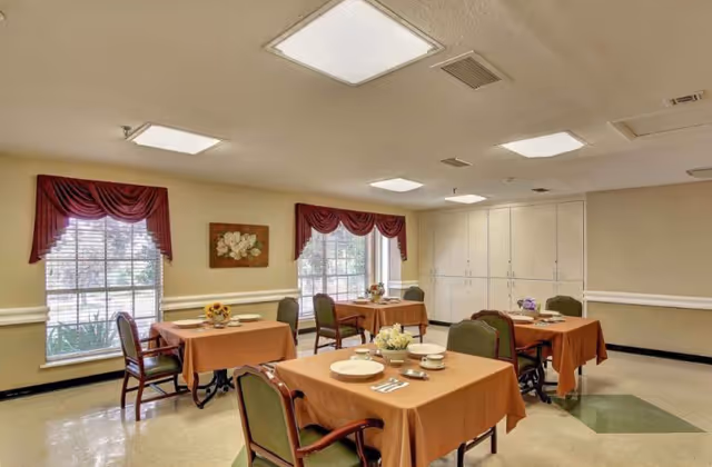 A dining room with four tables covered in brown tablecloths, each set with plates, cups, and utensils. The room has large windows with red valance curtains, beige walls, and ceiling lights. There are green cushioned chairs around the tables and a painting on the wall between the windows.