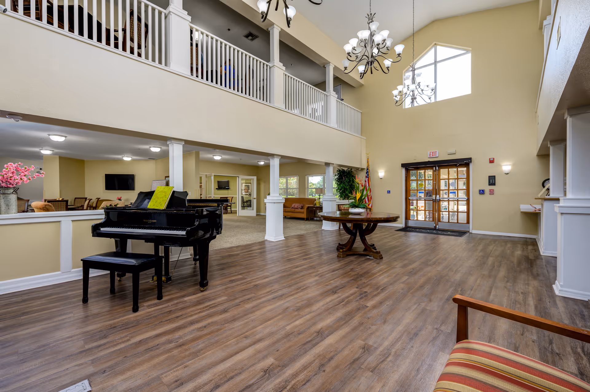 Spacious senior living facility common area with high ceilings, large windows, and wooden flooring. The room features a black grand piano with a bench, a round wooden table with a plant centerpiece, chandeliers, and seating areas with chairs and sofas. There is a balcony railing on the upper level and double glass doors at the entrance.