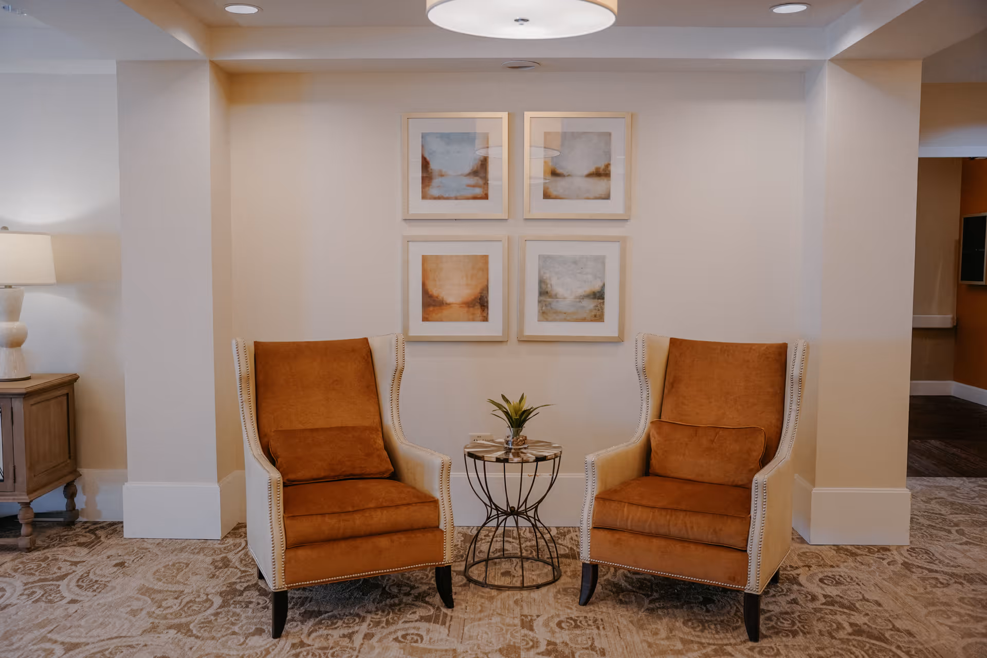 Two brown upholstered armchairs with matching cushions are positioned on either side of a small round metal table with a plant on top. Behind the chairs, there are four framed abstract art pieces arranged in a square on a beige wall. To the left, there is a wooden side table with a lamp. The floor is covered with a patterned carpet.