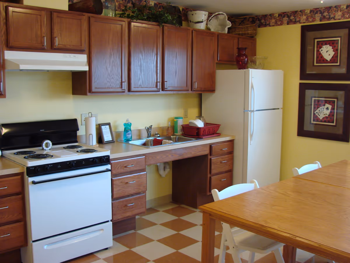 A bright kitchen with wooden cabinets, a white stove and refrigerator, a sink with dish rack, and a wooden dining table with chairs.