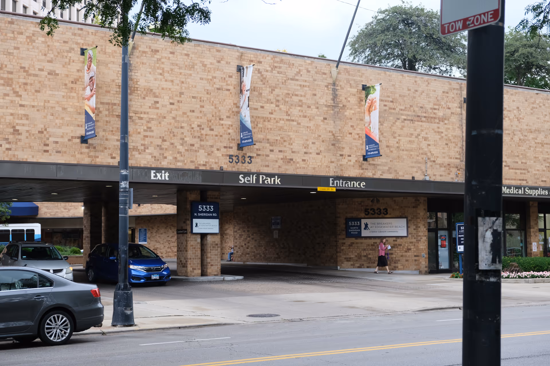 Brick-faced building front with a drive-through canopy labeled 'Exit Self Park Entrance' and address 5333, cars parked nearby and a pedestrian walking past.