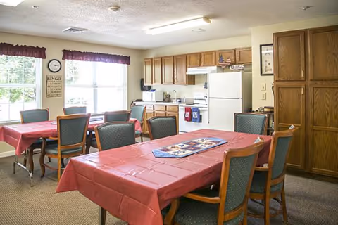 Communal dining room with tables draped in red tablecloths, green-upholstered chairs, and a small kitchen with oak cabinets and white appliances.