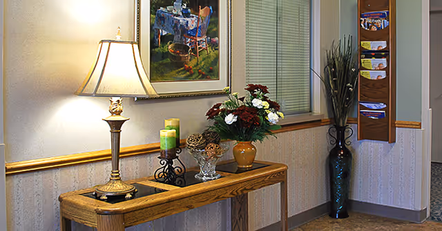 A warmly lit interior hallway corner featuring a wooden console table with a decorative lamp, candles, a glass bowl with decorative spheres, and a vase with red and white flowers. Above the table hangs a framed painting. To the right, there is a tall vase with long green plants and a wooden wall-mounted brochure holder with various pamphlets. A window with closed blinds is visible behind the table.