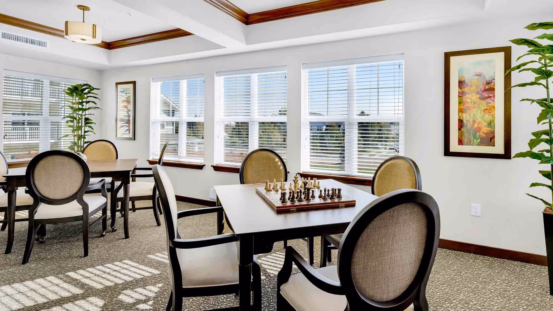 A bright and airy room with large windows letting in natural light. The room features multiple tables with cushioned chairs arranged around them. One table in the foreground has a chessboard set up for a game. The walls are decorated with framed artwork and there are tall potted plants adding greenery to the space. The ceiling has a tray design with crown molding and a modern light fixture.