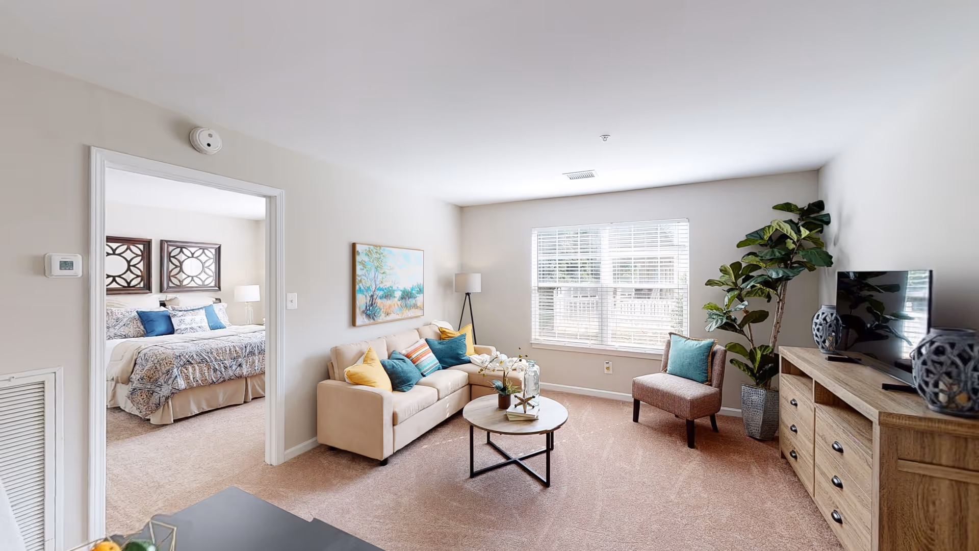 A bright living room with a beige sectional sofa adorned with colorful throw pillows, a round wooden coffee table with decorative items, a single upholstered chair with a teal pillow, a large potted plant, and a wooden TV stand with a flat-screen TV and decorative vases. A window with white blinds lets in natural light. Through an open doorway, a bedroom with a bed, patterned bedding, and two framed mirrors above the headboard is visible.