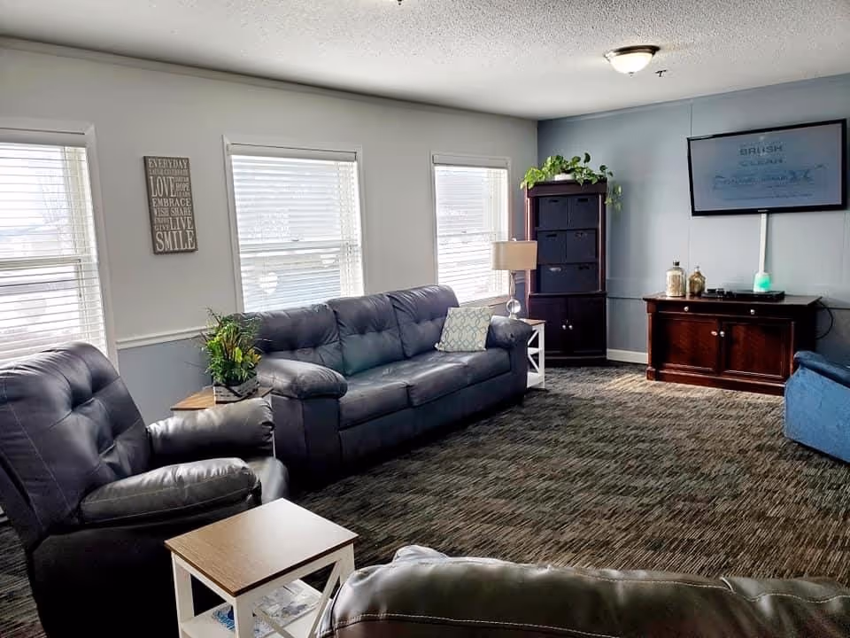A bright common living room with leather sofas and armchairs, side tables, plants, and a wall-mounted TV.