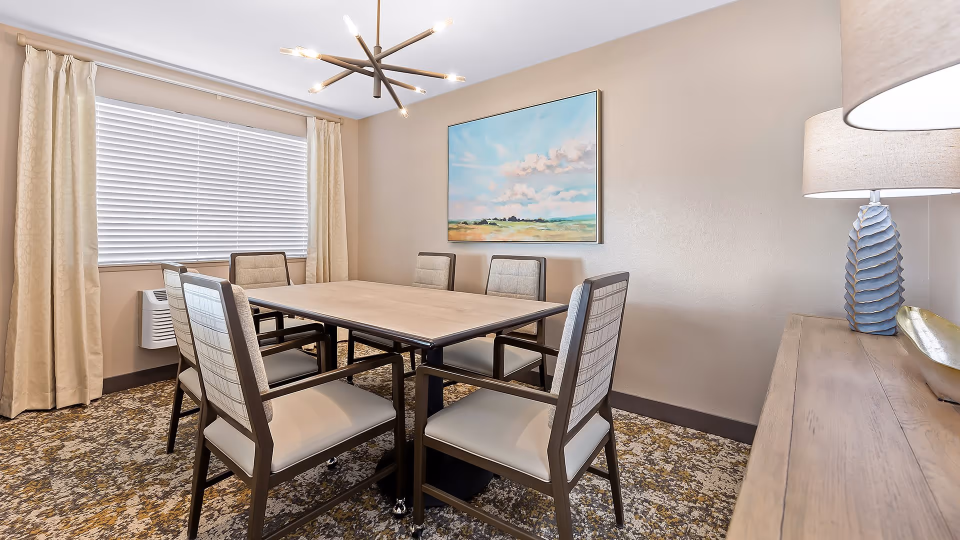 A dining room with a rectangular wooden table surrounded by six cushioned chairs. The room has beige walls, a large window with closed blinds and beige curtains, a modern chandelier, a landscape painting on the wall, and a wooden sideboard with a blue textured lamp and decorative bowl.
