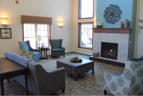 A cozy living room with a fireplace, two large windows, and several upholstered chairs and sofas arranged around a wooden coffee table. The fireplace has a decorative metal wall art above it and candles on the mantel. The room is well-lit with wall sconces and natural light from the windows.