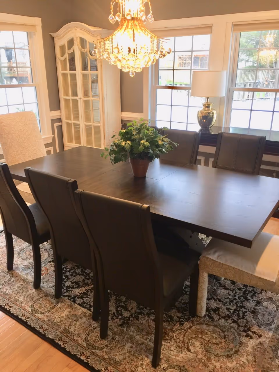 A dining room with a dark wooden table surrounded by six dark chairs and two light upholstered chairs at the ends. A potted green plant is centered on the table. The room features a large ornate chandelier hanging above the table, three large windows letting in natural light, a white display cabinet in the corner, and a table lamp on a sideboard beneath the windows. The floor has a patterned area rug.