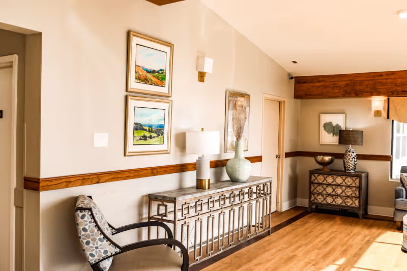 Sunlit sitting area with a decorative console table, lamp, artwork, and chairs in a senior living facility interior.