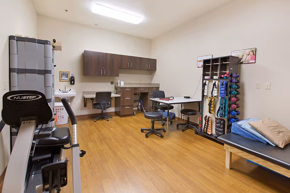 A therapy or exercise room with wooden flooring, a NuStep exercise machine, a table with chairs, a rack holding various exercise weights and bands, a therapy bed with pillows, and cabinets with a countertop and a sink in the background.