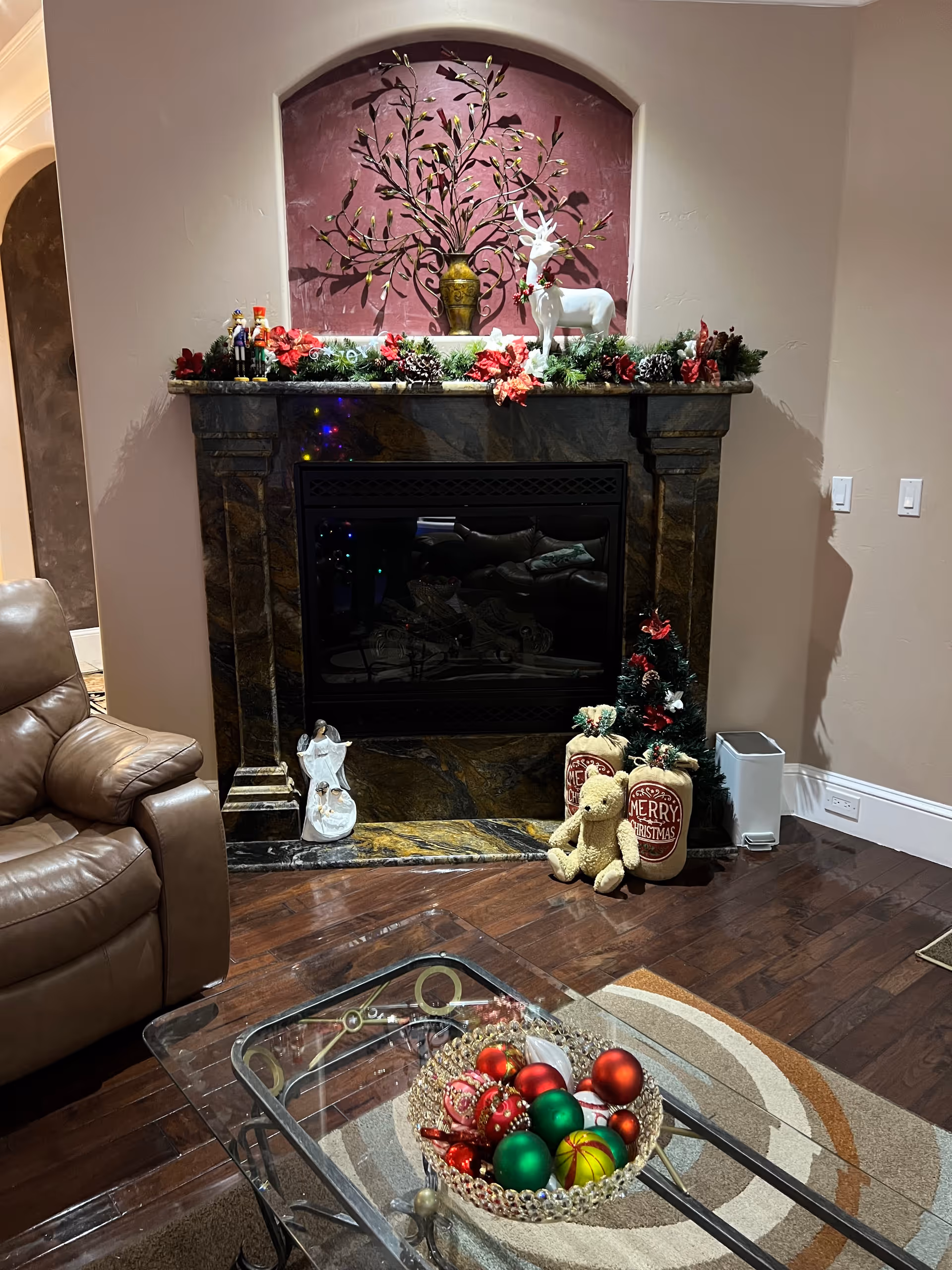 A cozy living room corner featuring a dark marble fireplace decorated with Christmas garlands, pine cones, and red flowers. On the mantel, there is a decorative vase with metal branches and a white reindeer figurine. Below the fireplace, there is a small Christmas tree, a teddy bear, and two Christmas-themed bags. To the left, part of a brown leather armchair is visible. In front, a glass coffee table holds a decorative bowl filled with red, green, and gold Christmas ornaments. The room has wooden flooring and a circular patterned rug.