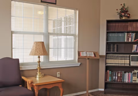 A cozy interior corner of a room with a window covered by white blinds, a wooden side table with a brass lamp, a purple upholstered chair, a wooden book stand holding an open book, and a tall bookshelf filled with various books and a small flower arrangement on top.