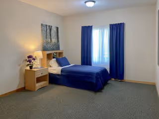 A simple bedroom with a single bed covered in blue bedding, a wooden nightstand with a lamp and a small vase of purple flowers, a window with blue curtains, and a framed picture of trees on the wall.