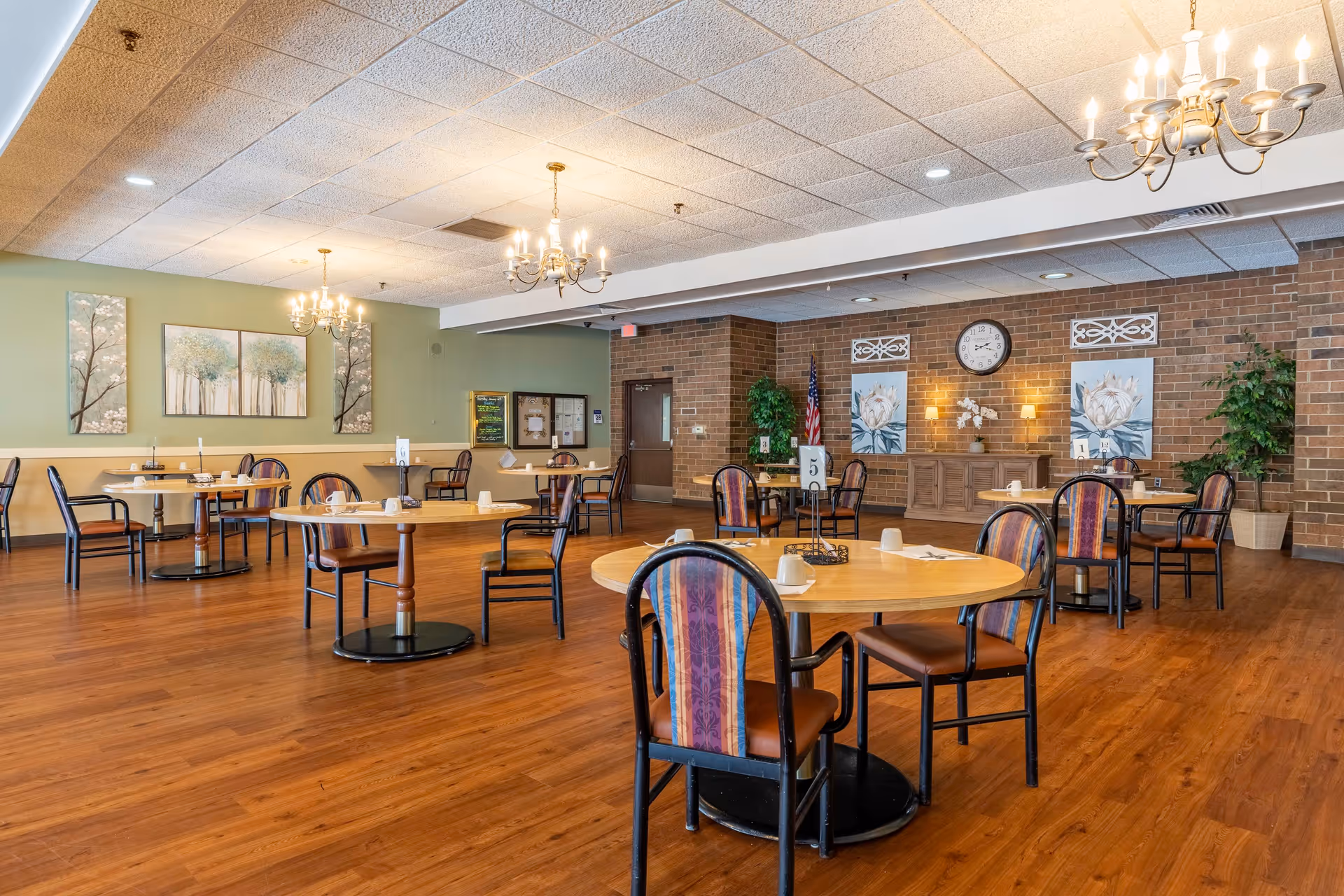 A spacious dining room with round wooden tables and chairs arranged neatly on a polished wooden floor. The walls are decorated with paintings of trees and flowers, and there are chandeliers hanging from the ceiling providing warm lighting. There are also potted plants and a clock on a brick accent wall in the background.
