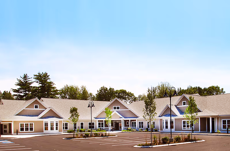 Exterior view of a single-story senior living facility building with multiple peaked roofs, large windows, and a spacious parking lot with young trees and lamp posts under a clear blue sky.