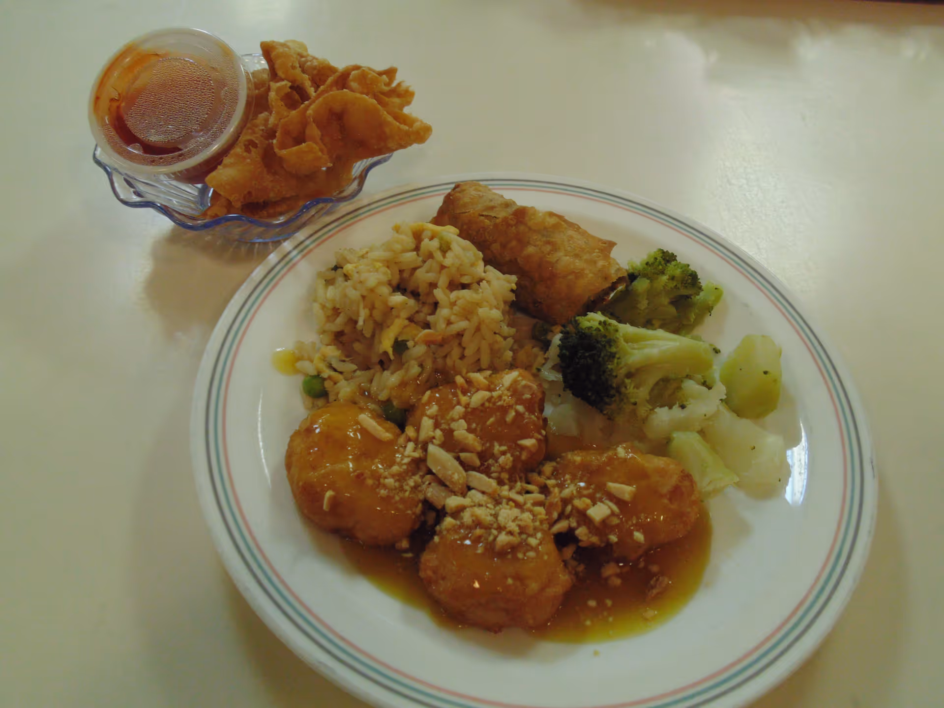 Plate of Asian-style food with rice, broccoli, an egg roll, sauced chicken pieces topped with chopped peanuts, and a small dish of fried wontons with dipping sauce on a table.