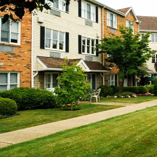 Exterior view of a multi-story residential building with brick and siding facade, surrounded by green shrubs, trees, and a well-maintained lawn with a sidewalk in front.