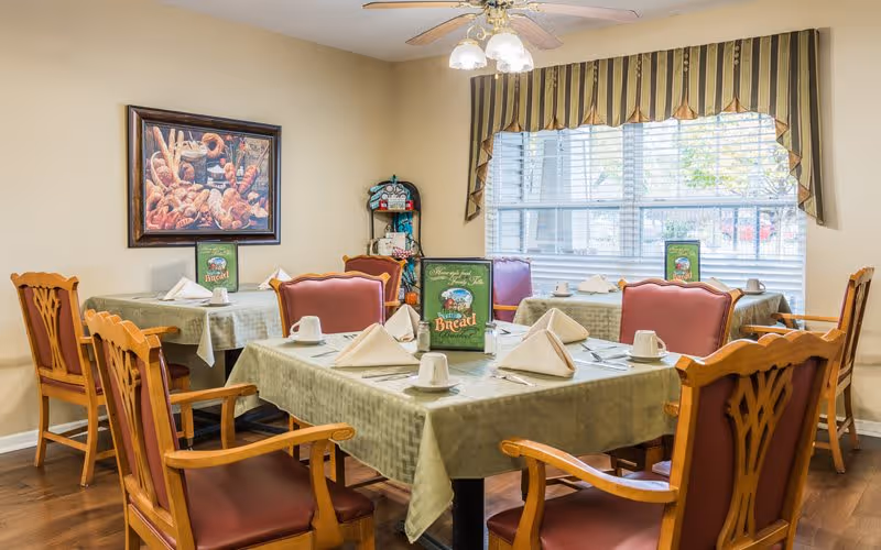 A dining room with tables covered in green tablecloths, each set with white napkins folded in a triangle, white cups, and silverware. The chairs have wooden frames with red cushioned seats and backs. A large window with striped valance curtains lets in natural light. A framed picture of bread hangs on the wall.