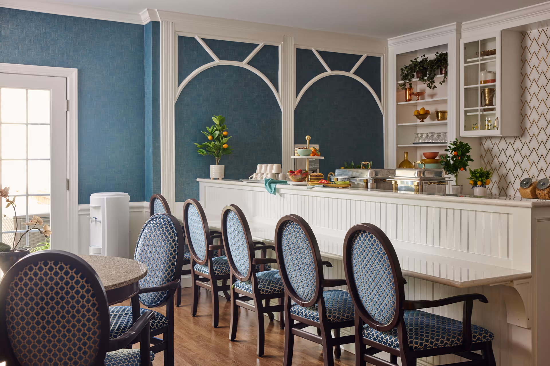 A dining area with a row of blue upholstered chairs with wooden frames lined up against a white counter. The counter has food trays and decorative plants on it. The walls are blue with white decorative molding, and there is a door with glass panels on the left side. Shelves with glassware and plants are visible behind the counter.