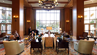 Spacious, elegant lounge with high ceilings and a large window, chandelier, and arranged sofas and chairs with several people seated.