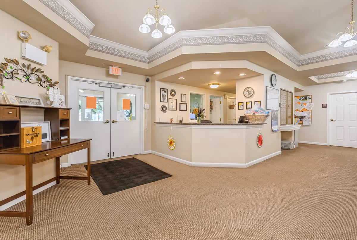 Reception area of a senior living facility with a front desk, double doors with windows, a wooden suggestion box on a small table, wall decorations, and a bulletin board. The area is carpeted and well-lit with ceiling lights.