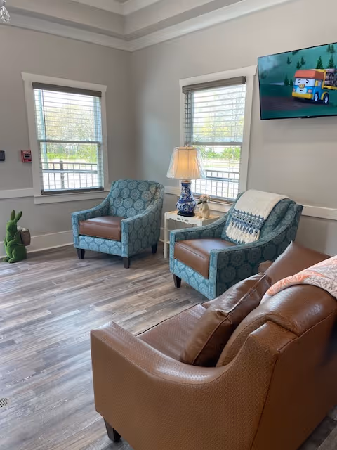 A cozy living room area with two patterned blue armchairs and one brown leather armchair arranged around a small white side table with a decorative lamp and figurine. Two windows with blinds let in natural light, and a flat-screen TV is mounted on the wall showing a colorful animated scene. The floor is wood with a light finish.