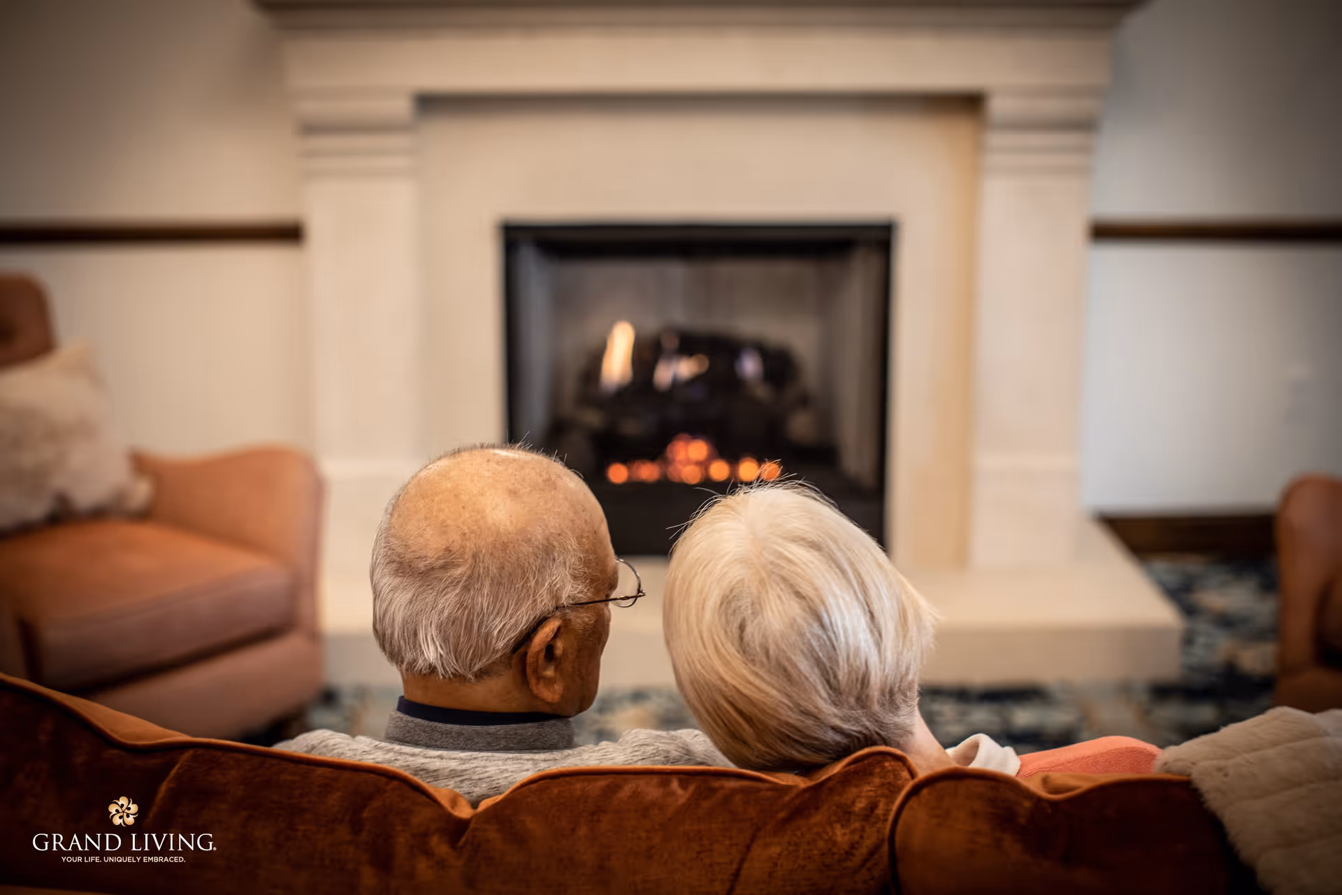 An elderly couple sitting closely together on a brown couch, facing a lit fireplace in a cozy living room setting.