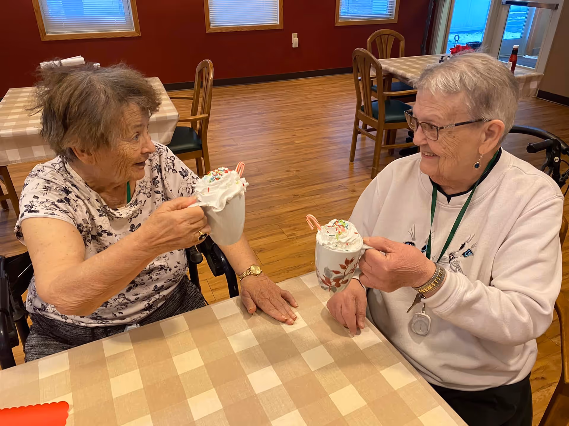 Two elderly women sit at a dining table smiling and clinking mugs topped with whipped cream and candy canes.