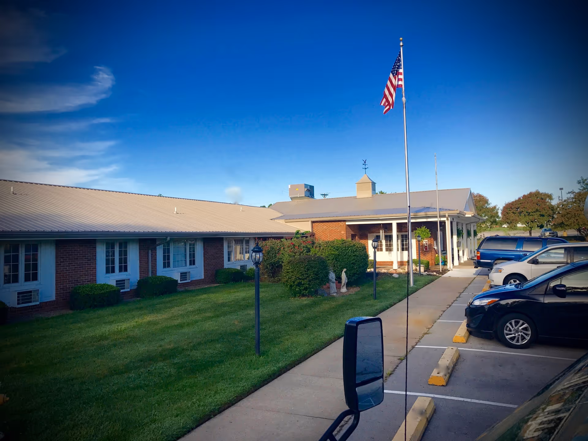 Front exterior of a single-story brick building with an American flag, lawn and parked cars.