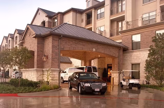 Entrance of a multi-story senior living facility with a covered driveway. A black car, a white van, and a golf cart are parked near the entrance. The building features brick and stone exterior walls with multiple windows and balconies. There are small trees and landscaping around the driveway.