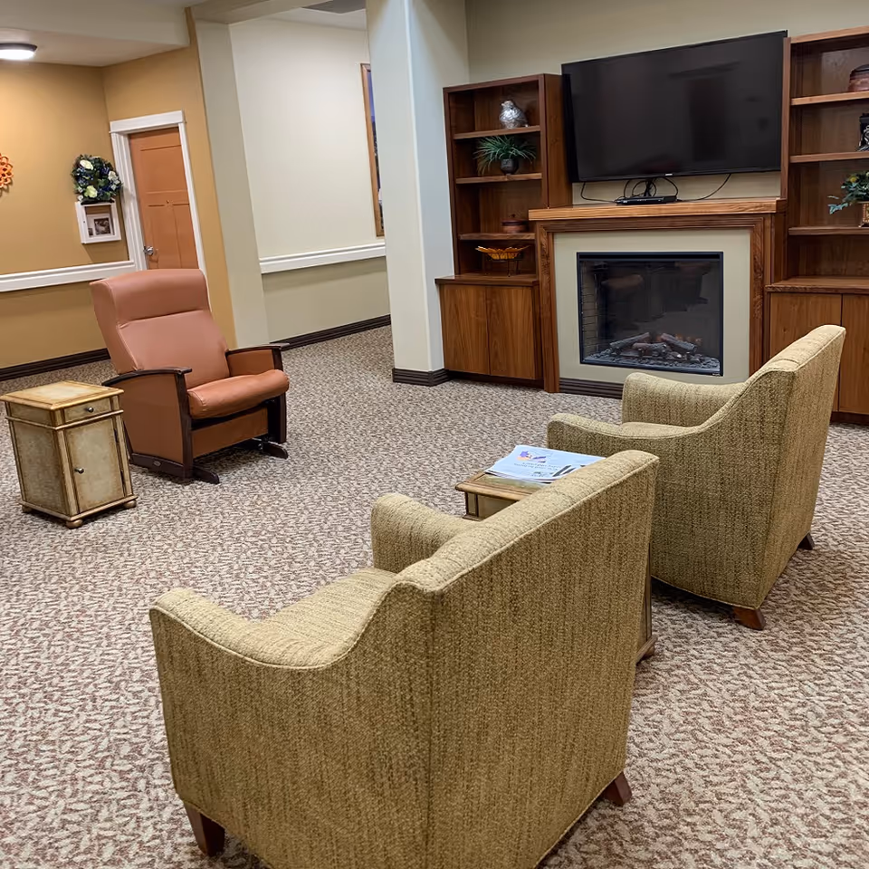 A cozy living room area in a memory care facility featuring two beige armchairs facing a small wooden table with papers on it. Across from them is a brown recliner chair next to a small wooden cabinet. The room has a carpeted floor, a wall-mounted flat-screen TV above an electric fireplace, and wooden built-in shelves with decorative items.