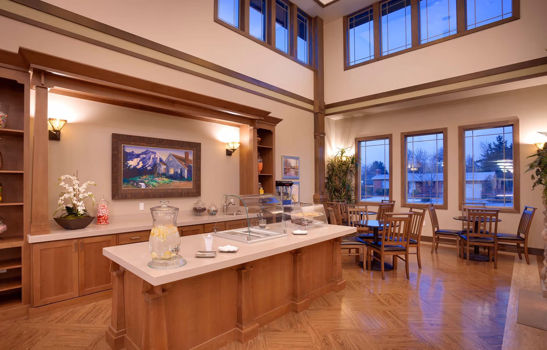 Spacious, well-lit dining/common area with a wooden central service island, tables and chairs, large windows and high ceilings.