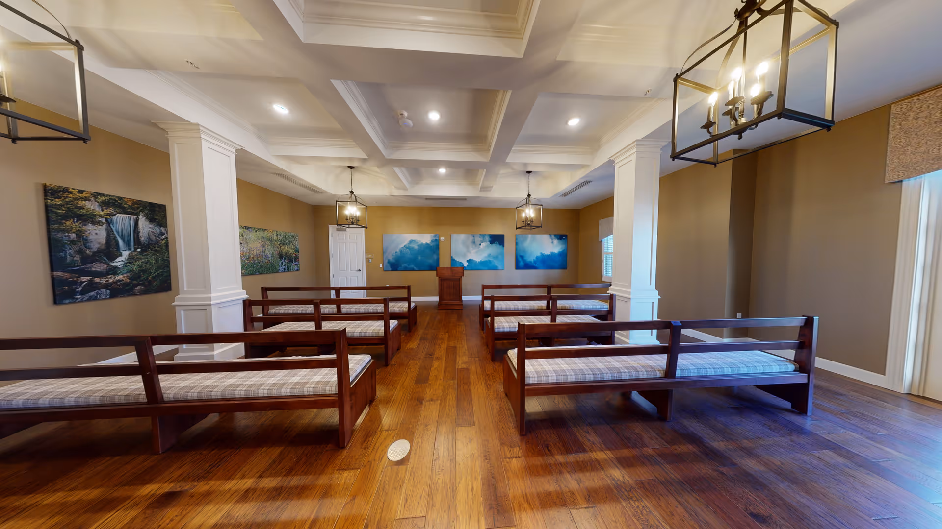 Interior view of a room with wooden benches arranged in rows facing a wooden podium. The room has hardwood flooring, beige walls, white columns, and ceiling lights with decorative fixtures. There are landscape paintings on the walls and windows with blinds on the right side.