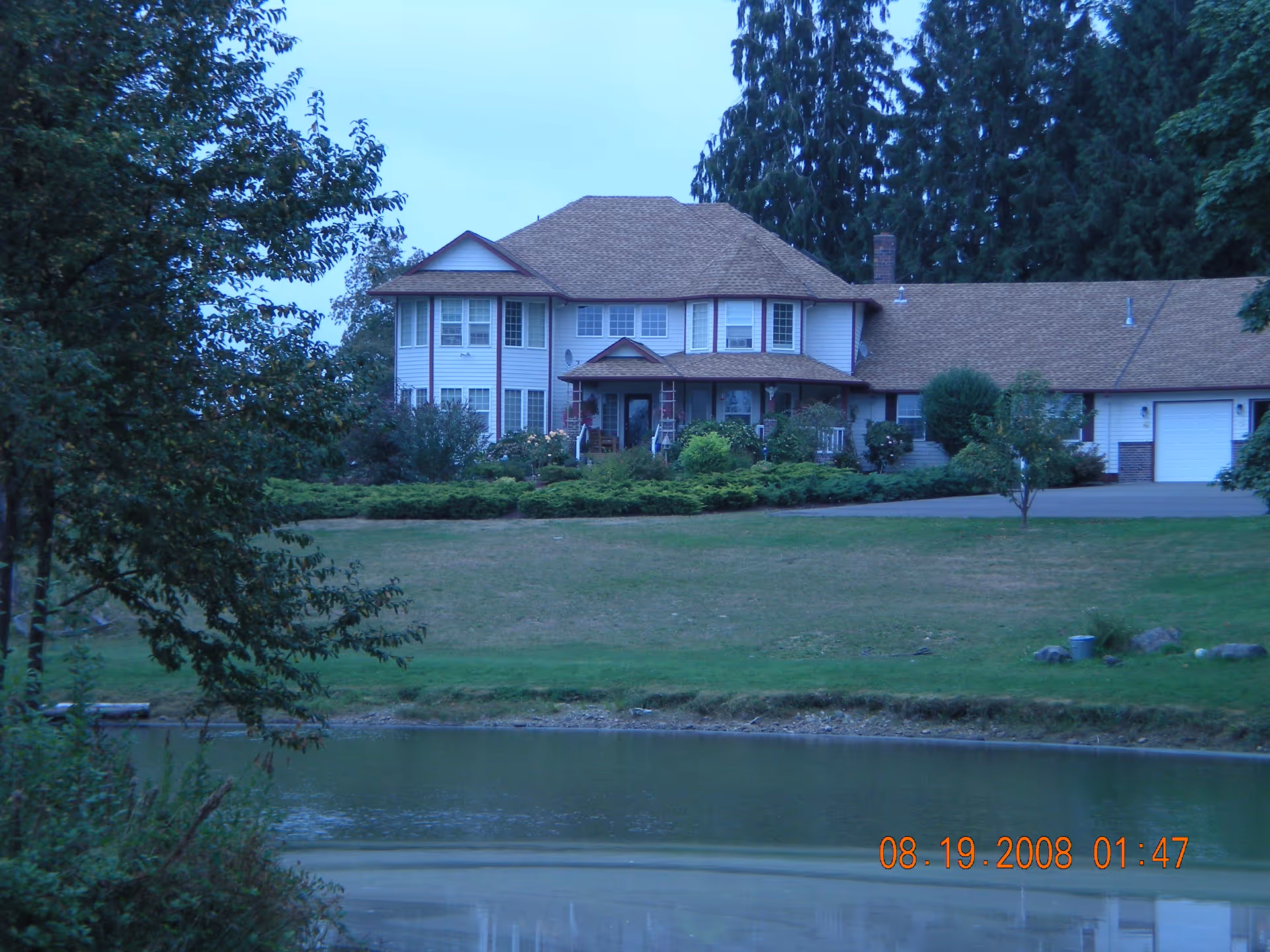 A large two-story house with a brown roof and white exterior, surrounded by trees and bushes, with a pond in the foreground and a grassy lawn between the pond and the house.