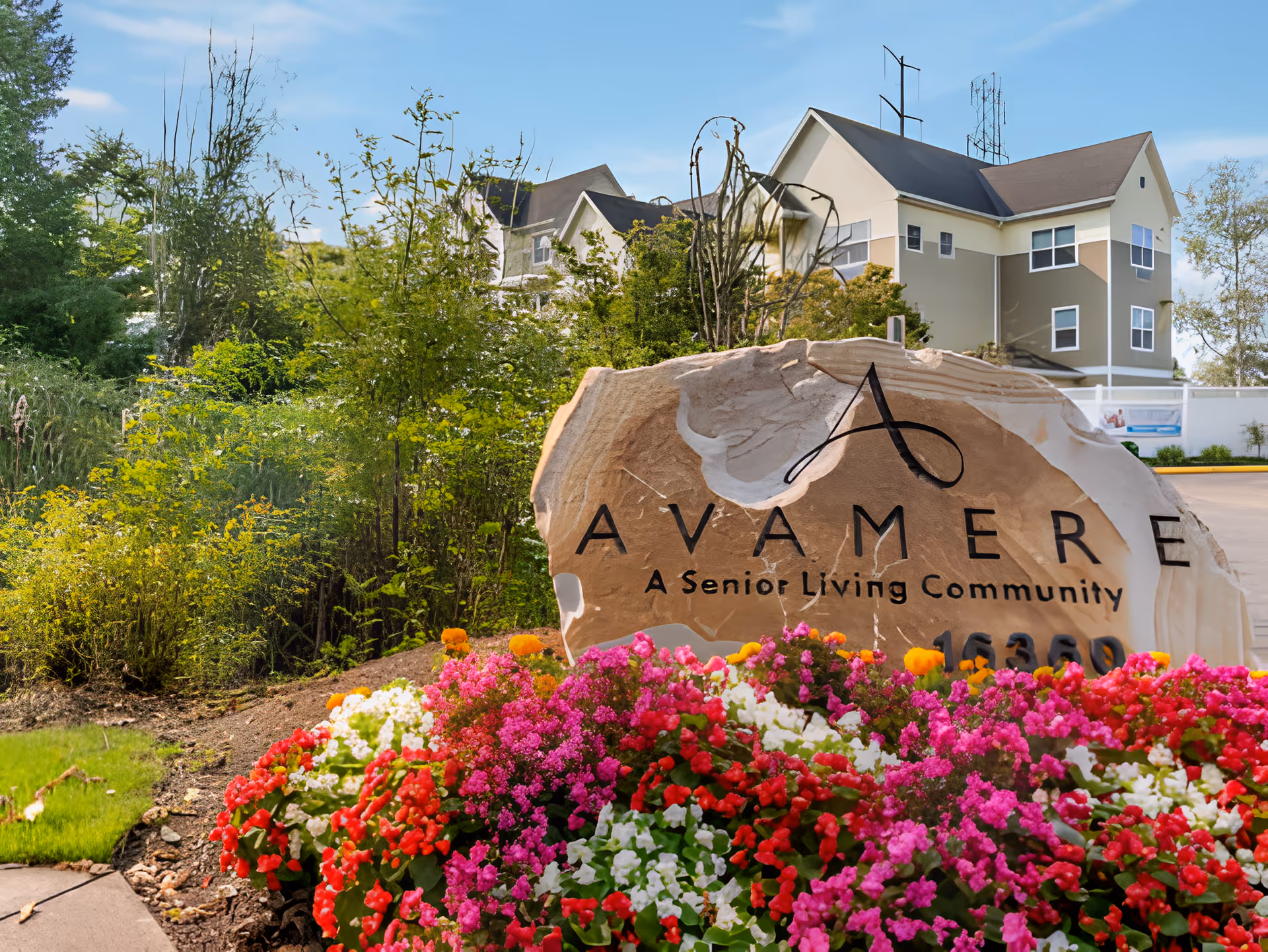A large stone sign with the text 'Avamere A Senior Living Community' surrounded by colorful flowers in front of a senior living facility building with trees and a clear blue sky in the background.