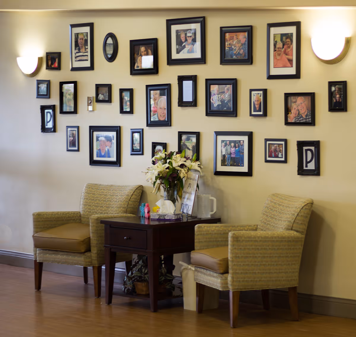 Two cushioned armchairs with a wooden side table between them, holding a vase of flowers and some small items. Behind the chairs is a wall decorated with multiple framed photographs of people, arranged in a grid pattern. The wall is illuminated by two wall-mounted lights on either side.
