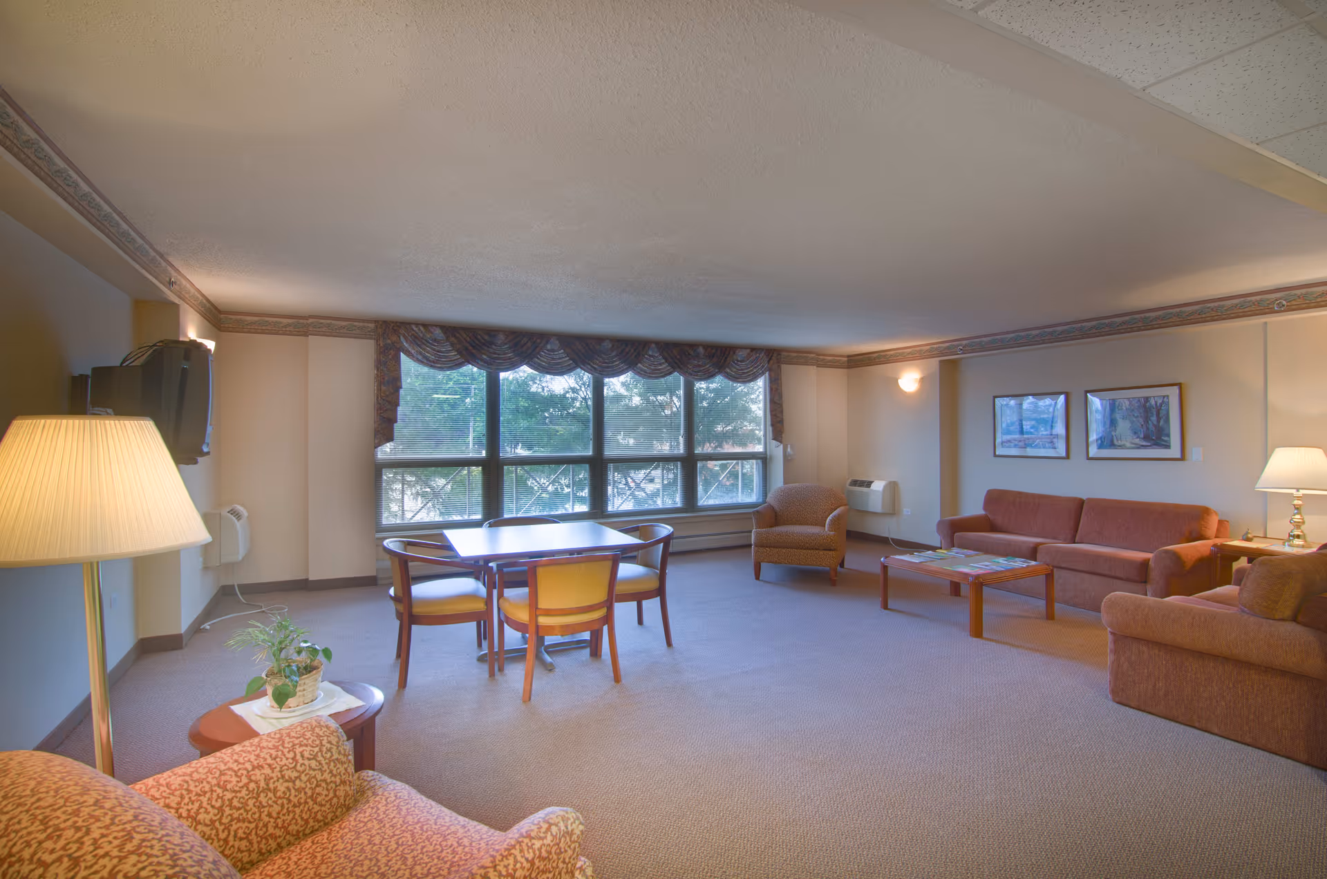 A spacious living room with large windows covered by valance curtains, allowing natural light to fill the room. The room is furnished with a sofa, two armchairs, a coffee table, a small dining table with four chairs, a floor lamp, and a wall-mounted television. The walls are decorated with two framed pictures, and there is a small side table with a lamp and a plant.