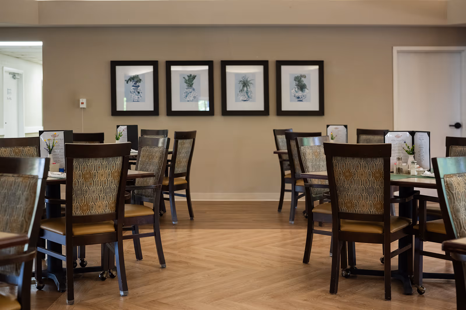 A dining room with several wooden tables and chairs arranged neatly. Each table has a menu and small decorative plants. The walls are beige with four framed botanical prints hanging in a row. The floor has a light wood pattern.