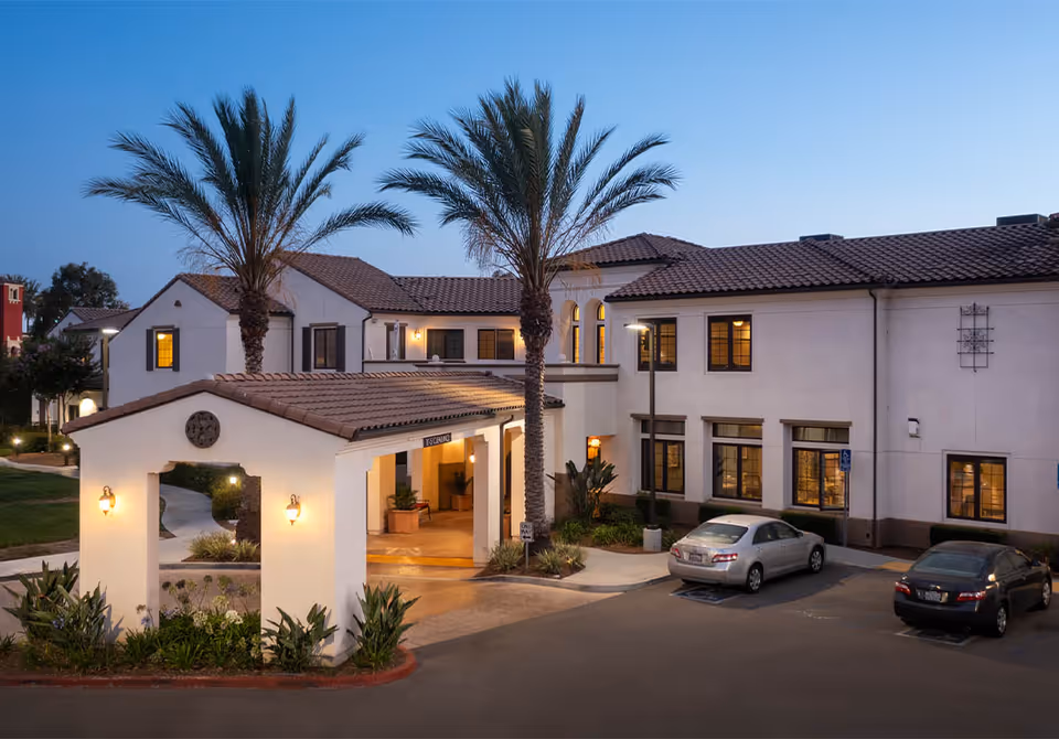 Exterior front entrance of a Mediterranean-style senior living building with a lit porte-cochère, palm trees, and parked cars at dusk.