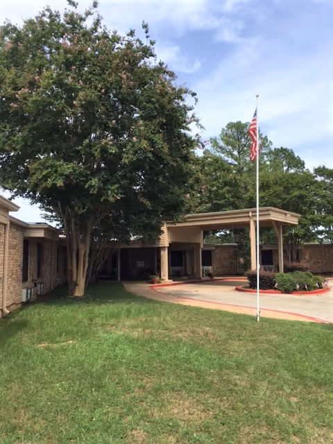 Exterior view of a single-story brick building with a covered entrance and an American flag on a flagpole in front. There is a large tree to the left and green grass surrounding the driveway.