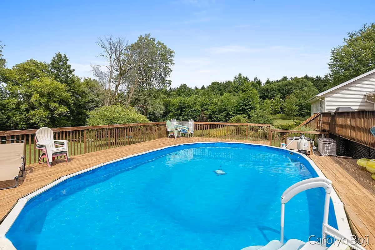 Outdoor swimming pool surrounded by a wooden deck with several plastic chairs and lounge chairs. The pool area is adjacent to a white building and is surrounded by green trees and bushes under a clear blue sky.