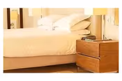 A neatly made bed with white pillows and sheets next to a wooden nightstand with two drawers. A lamp and a telephone are placed on the nightstand.