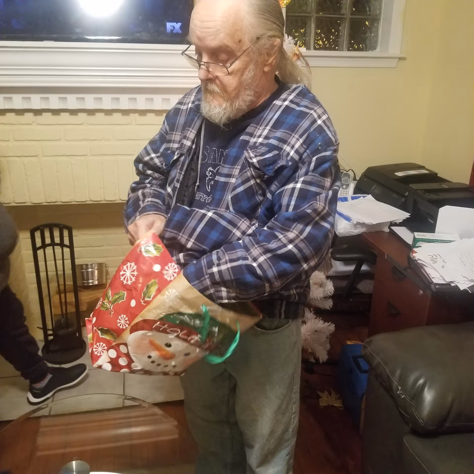 An elderly man with glasses and a beard wearing a blue plaid shirt is standing indoors, holding a festive holiday gift bag with a snowman design. Behind him is a fireplace with a TV mounted above it, a desk cluttered with papers and office equipment, and part of a leather chair is visible.