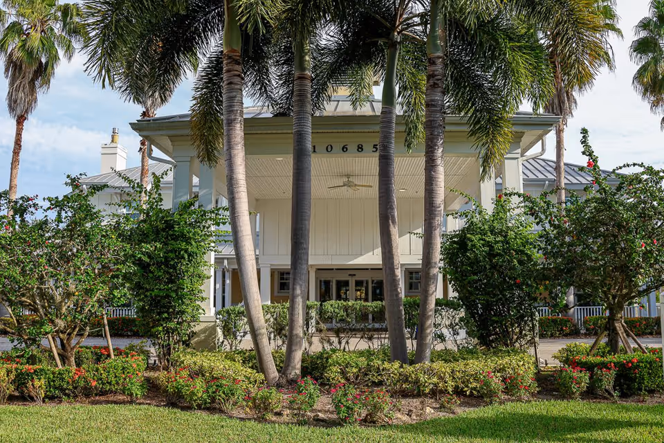 Front exterior view of The Brennity at Tradition Senior Living building with palm trees and landscaped bushes in the foreground under a partly cloudy sky.