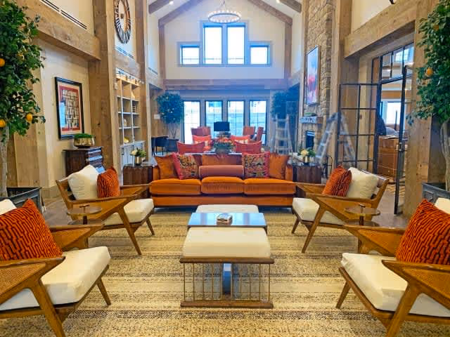 A spacious and well-lit living room with high vaulted ceilings and large windows. The room features a burnt orange sofa with colorful cushions, four wooden armchairs with white cushions and orange pillows, a central coffee table, and a patterned rug. There are plants on either side of the room, a large wall clock, and a fireplace with a painting above it in the background.