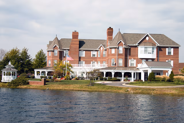 Large brick senior living facility building with multiple chimneys and a wraparound porch, situated beside a body of water with a small gazebo on the left side and landscaped greenery around.