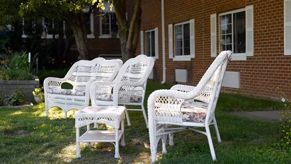 White wicker outdoor furniture including two armchairs, a loveseat, and a small table arranged on a grassy lawn in front of a brick building with white-framed windows.