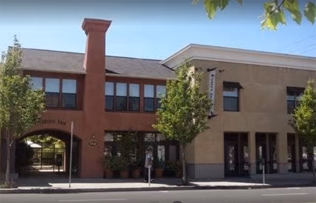 Street-facing view of the two-story Elders Inn building with a central arched entrance, storefront windows, and trees along the sidewalk.